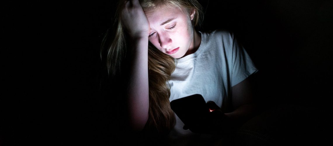 Upset girl sitting in the dark while using her smartphone. The l